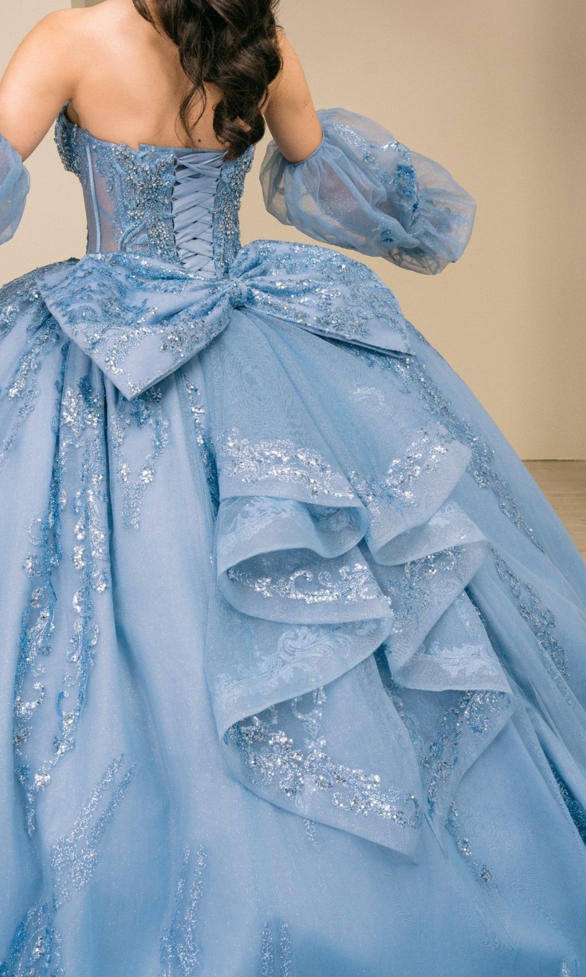 A person wearing the Cinderella Couture Quinceanera Dress 8565JQ, a strapless blue ball gown featuring a lace-embellished corset bodice, decorative ruffles and a large bow on the back, and removable puff sleeves for versatile styling.